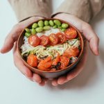 Close-up of hands holding a salmon poke bowl with cherry tomatoes, edamame, and rice.